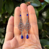 A pair of silver filigree dangle earrings with small blue beads and teardrop-shaped blue gemstones, held in a person's hand against a background of green leaves.