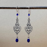 A pair of silver filigree dangle earrings with deep blue teardrop beads, hanging from a metal rod against a neutral beige background.