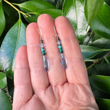 Blue earrings held in a hand with leaves in the background. 