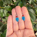 A pair of dangling earrings with light blue teardrop-shaped beads and small red beads, held between fingers against a background of green leaves.