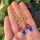 Gold earrings held in a hand with blue glass and red crystal.