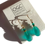 A pair of dangle earrings with teardrop-shaped teal beads, small faceted green beads, and gold-tone hooks, displayed on a textured white card labeled 'OGC OWEN GLASS COLLECTION'.