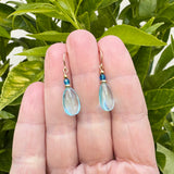Teardrop-shaped earrings with blue and gold accents held between fingers against a green leafy background