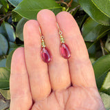 Pink glass drop earrings with peridot crystal top beads. Gold trim, made in the USA. Earrings are held in hand with leafy background.