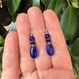 A pair of cobalt blue glass teardrop dangle earrings with silver and black spacer beads, held in a person's hand with green leaves in the background.