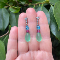 Pair of earrings with green teardrop beads and colorful beads on a hand against a leafy background