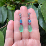 Pair of earrings with green teardrop beads and colorful beads on a hand against a leafy background