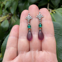 Pair of earrings with purple teardrop beads and silver floral tops held between fingers against a green leafy background.