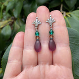Pair of earrings with purple teardrop beads and silver floral tops held between fingers against a green leafy background.