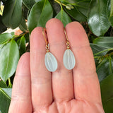 Gold earrings with frosted teardrop-shaped stones held between fingers against a green leafy background