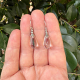 A pair of clear teardrop-shaped glass dangle earrings with small silver beads and hooks, held between fingers against a background of green leaves.