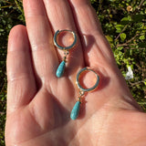 A pair of gold huggie hoop earrings with textured turquoise stone teardrop charms, shown resting on an open hand with a blurred green garden background.