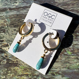 A pair of gold-tone huggie hoop earrings with dangling turquoise-blue teardrop glass beads, displayed on a white branded card that reads 'OGC Owen Glass Collection.' The card is photographed on a textured stone-like surface in bright sunlight, casting strong shadows.