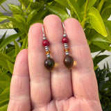 Green, red and orange glass earrings held in a hand with a leafy background.