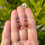 Red and blue earrings held in a hand with a leafy green background.