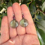 A pair of handmade dangle earrings held in a person's hand, featuring round olive-green cabochon stones and small purple beads, set against a background of green leaves.