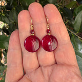 A pair of red round drop earrings with gold hooks and small faceted beads, held in a person's hand with green leaves in the background.